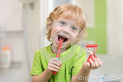 Happy child boy brushing teeth near mirror in bathroom. He is monitoring lasting of cleaning action with hourglass. Stock Photo