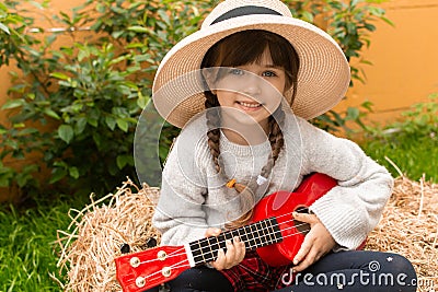 Happy kid girl in straw hat with ukulele having fun on summer field. Stock Photo