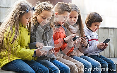 Happy kids sitting on bench with mobile devices Stock Photo