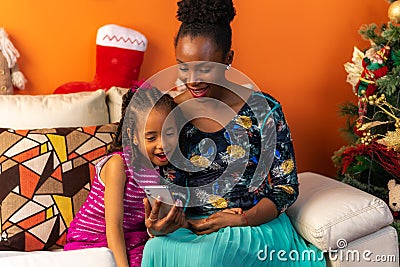Happy Latin Afro-descendant Colombian mother and daughter sitting on the sofa next to the Christmas tree checking the cell phone Stock Photo