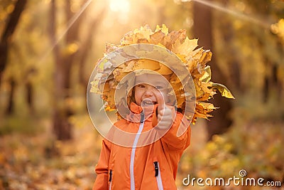 Happy laughing toddler child in crown made of autumn fallen leaves showing thumb up. Happiness and seasonal activities concept Stock Photo