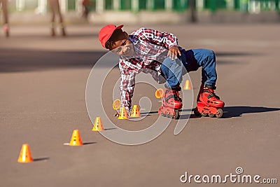 Happy little african american boy on rollers skates in summer park at asphalt road among training cones . Roller kid Stock Photo