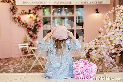 Happy little curly hair girl in dress and beret with bouquet of peonies sitting in street vintage cafe, view from back. Childhood Stock Photo