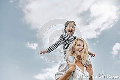 Happy little funny daughter on a piggy back ride with her happy mother on the sky background. Loving woman and her little girl Stock Photo