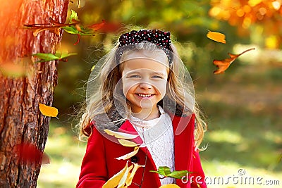 A happy little girl in a bright red coat and a white knitted sweater with a polka-dot headband plays in autumn park. Childhood. Stock Photo