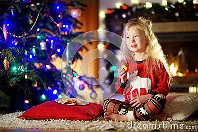 Happy little girl having milk and cookies by a fireplace in a cozy dark living room on Christmas eve Stock Photo