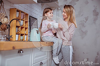 Happy little girl and her beautiful young mother have breakfast together in a white kitchen. They hug and drink tea. Maternal care Stock Photo