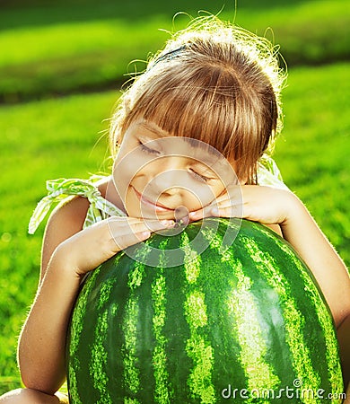 Happy little girl is hugging huge watermelon sitting on the green grass in summertime. Stock Photo