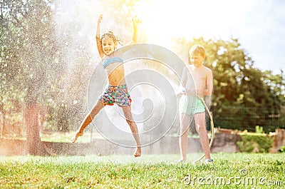 Happy little girl jumps under water, when brother pours her from garden hose. Hot summer days activity Stock Photo