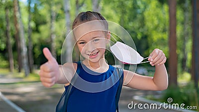 Happy little girl takes off the mask and shows thumb outdoors. Joyful smiling schoolgirl with a backpack pushes the mask Stock Photo
