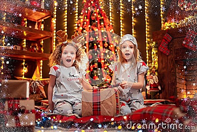 Happy little girls wearing Christmas pajamas open gift box by a fireplace in a cozy dark living room on Christmas eve. Stock Photo