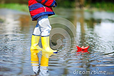 Happy little kid boy in yellow rain boots playing with paper ship boat by huge puddle on spring or autumn day Stock Photo