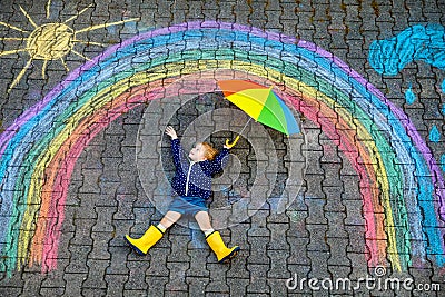 Happy little toddler girl in rubber boots with rainbow sun and clouds with rain painted with colorful chalks on ground Stock Photo