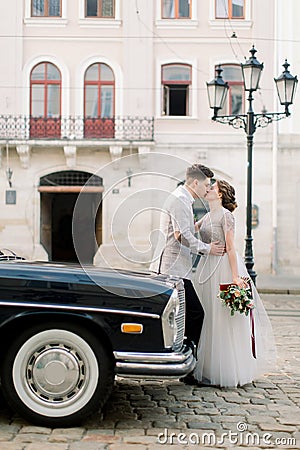 Happy luxury wedding couple kissing and embracing near black retro car in old city center, ancient buildings on the Stock Photo