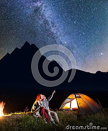 Happy male tourist showing his female at the stars in the night sky. Couple sitting near the glowing tent and campfire Stock Photo