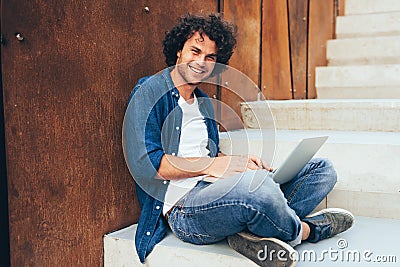 Happy man with curly hair using laptop for chatting online with friends, browsing on Internet on the city. Handsome male smiling Stock Photo