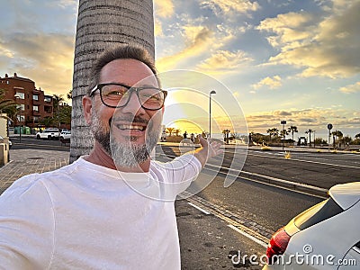 Happy man taking portrait selfie picture against a colorful sunset on the street with car and roads. Travel and summer vacation Stock Photo
