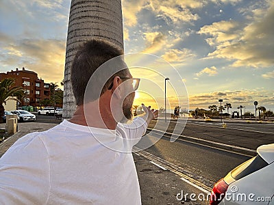 Happy man taking portrait selfie picture against a colorful sunset on the street with car and roads. Travel and summer vacation Stock Photo