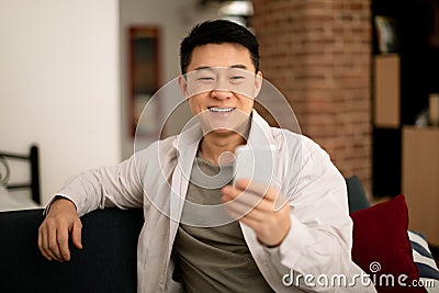 Happy mature asian man in casual watching video or chatting on smartphone, resting on sofa in living room interior Stock Photo