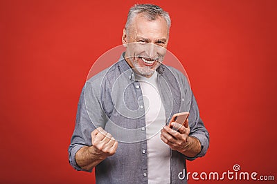 Happy mature senior man in casual using smartphone and making winner gesture isolated over red background. Using phone Stock Photo