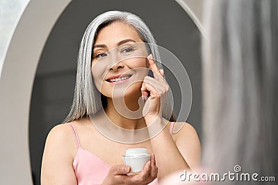 Happy middle aged mature asian woman looking at mirror applying antiage cream. Stock Photo