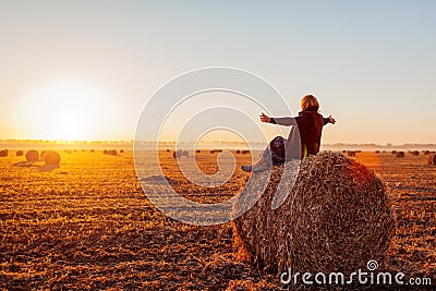 Happy middle-aged woman sitting on haystack in autumn field and feeling free with arms opened Stock Photo