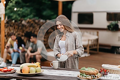 Happy millennial Caucasian woman pouring tea, making breakfast near RV, camping with her diverse friends outdoors Stock Photo