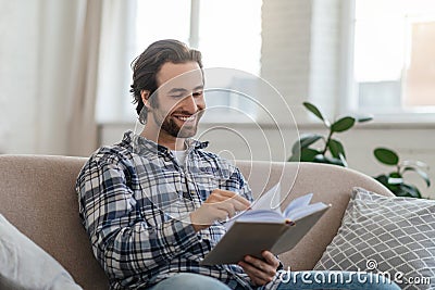 Happy millennial handsome caucasian guy with stubble sits on sofa reads book in living room interior Stock Photo