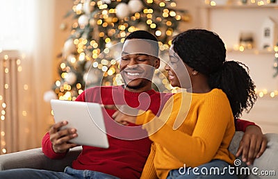 Happy millennial lady points finger at tablet and shows to guy something interesting in interior with tree and garlands Stock Photo