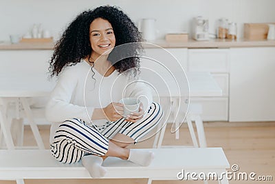 Happy morning concept. Photo of cheerful curly African American woman sits in lotus pose at white bench, sips tasty aromatic drink Stock Photo