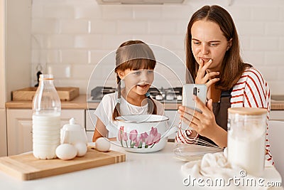 Happy mother baking together with her daughter, sitting at table in the kitchen and finding recipe in the internet via mobile Stock Photo