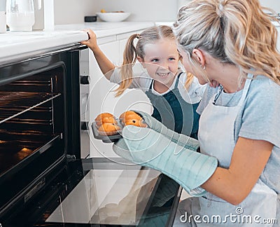 Happy mother and daughter removing muffins from the oven. Little girl looking at her parent holding a tray of muffins Stock Photo