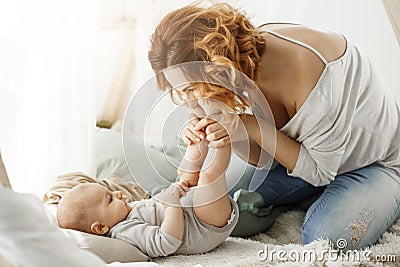 Happy mother playing with newborn baby kissing little legs spending best maternity moments in cozy bedroom. Warm family Stock Photo