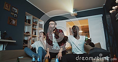 Happy multiethnic male friends shout and cheer watching sports game together at home on TV using projector slow motion. Stock Photo