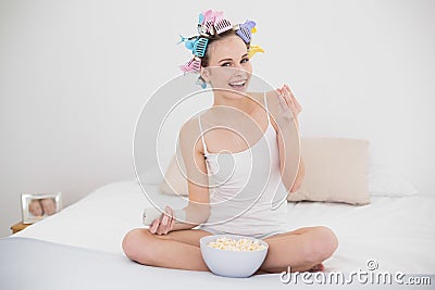 Happy natural brown haired woman in hair curlers watching tv while eating popcorn Stock Photo