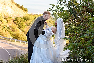 Happy newlyweds kiss against the backdrop of a beautiful mountain landscape, the girl holds the guy by the tie Stock Photo