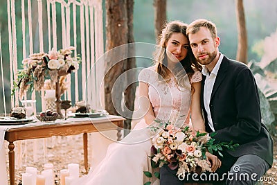 Happy newlyweds look on a photographer. Man and woman in festive clothes sit on the stones near the wedding decoration Stock Photo