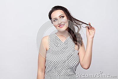 Happy optimistic beautiful young brunette woman with makeup and striped dress standing touching hair, dreaming and looking away Stock Photo