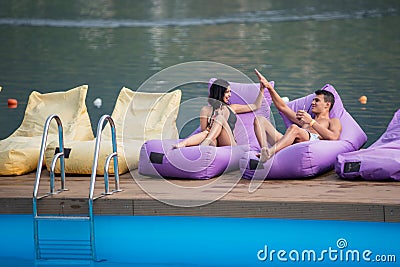 Happy pair relaxing with cocktail and giving high five to each other on cushioned loungers by swimming pool Stock Photo