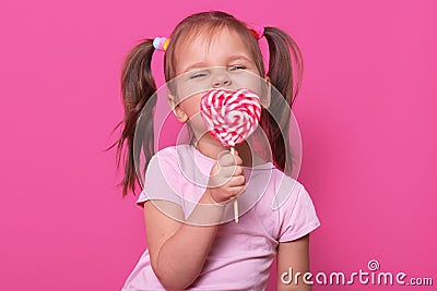 Happy playful cute girl wears rose t hirt, stands isolated over pink background, holds bright lollipop in hand, eating it. Happy Stock Photo