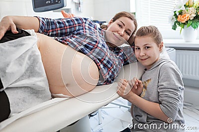 Happy pre teen daughter sitting near her pregnant mother with large belly in an ultrasonography room Stock Photo