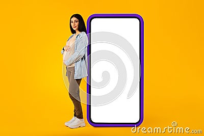 Happy pregnant woman leaning at big smartphone with blank screen and embracing her belly, advertising pregnancy app Stock Photo