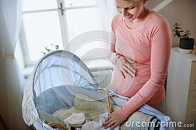 Happy pregnant woman looking at little clothes for her baby in cradle. Stock Photo