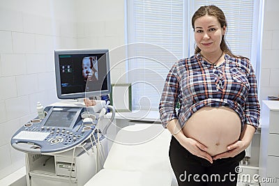 Happy pregnant woman standing in consulting room after ultrasonography near ultrasound scan with fetus 3d image on monitor screen Stock Photo