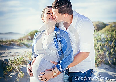 Happy pregnant young couple interacting, standing outdoors kissing, beach scene Stock Photo