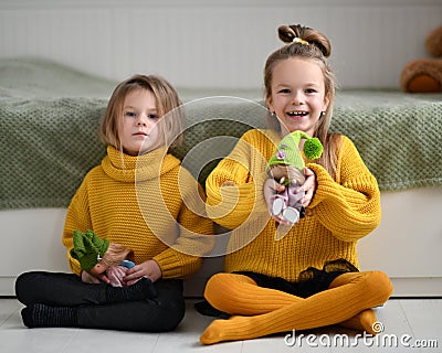 Happy pretty small girls sisters children in warm yellow knitted home sweaters sitting on floor and playing with dolls Stock Photo