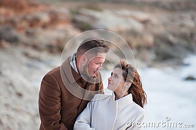 Happy pretty young family beautiful caucasian man in coat and scarf hugging smiling woman looking at each other with love on ocean Stock Photo