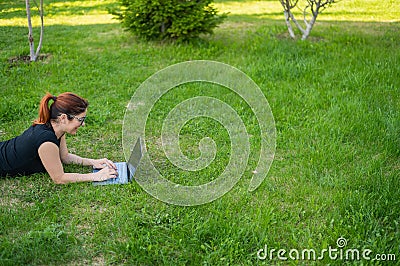 Happy red-haired girl in glasses lies on the lawn in the park and types on the laptop keyboard. Young caucasian female Stock Photo