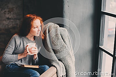 Happy redhead woman relaxing at home in cozy winter or autumn weekend with book and cup of hot tea, sitting in soft chair Stock Photo