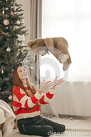 Happy redhead woman in sweater with toy bear under Christmas tree with lights. New Year holidays. Christmas Stock Photo